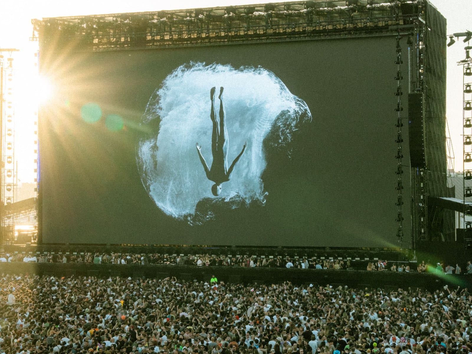An outdoor festival crowd at golden hour facing a giant LED wall carrying a single figure suspended in light