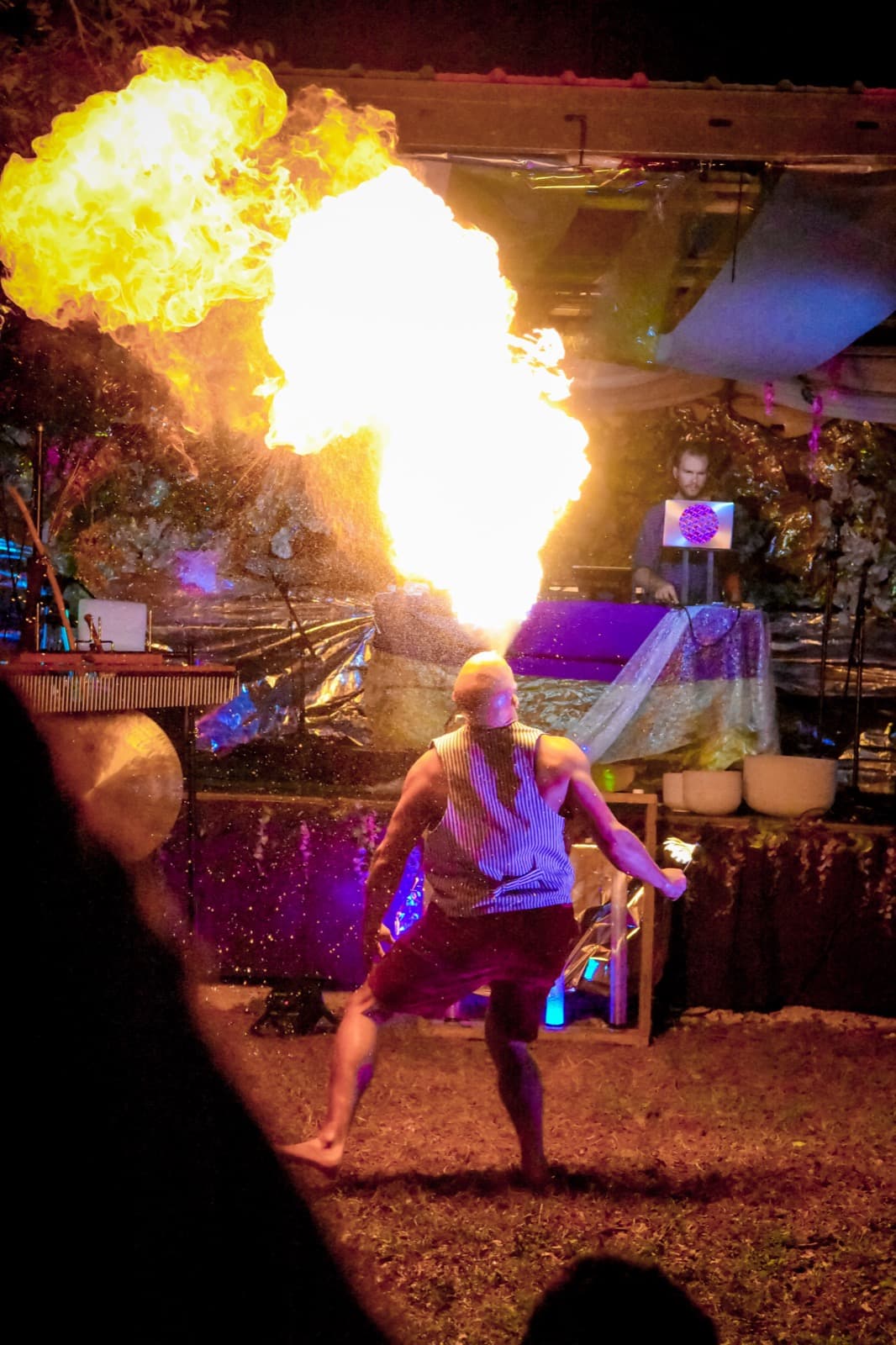 A fire breather sends a plume of flame over the crowd, a DJ behind